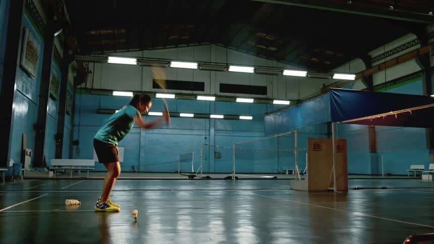 At an indoor badminton court an asian woman wearing shorts tries to hit a shuttlecock with her racket misses throws the racket down and walks away