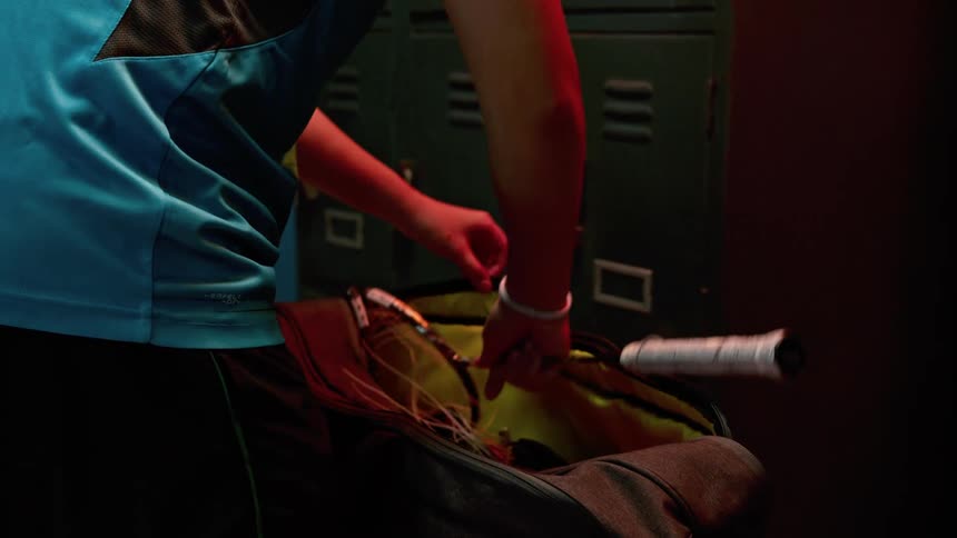 A girl in a locker room placing a broken racket in a gym bag with lockers and locks in the background