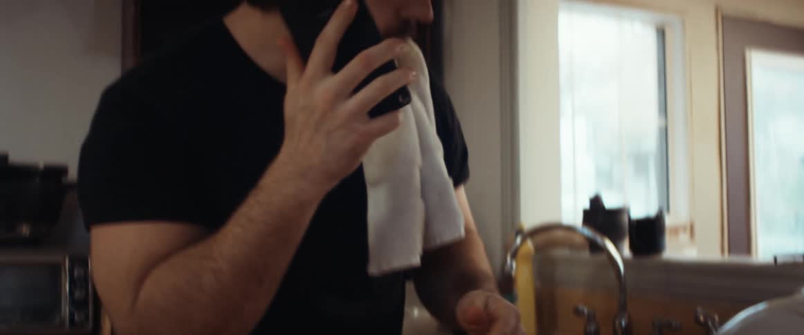 Man with a beard and a black t shirt does the dishes and answers incoming call in the kitchen