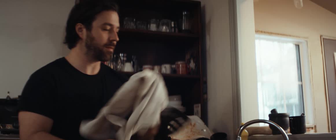 A man washing dishes at home looks over his shoulder then picks up a smart phone from off of a counter