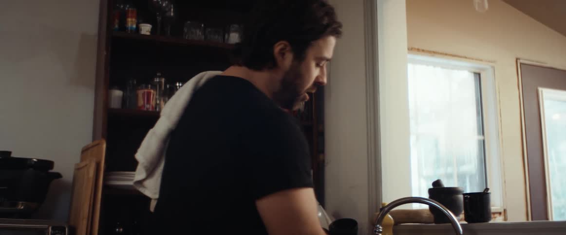 Man with a beard and a black t shirt does the dishes and looks at his smartphone in the kitchen