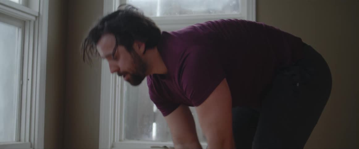 Muscular man with a beard and a t shirt lifts weights next to windows in a room