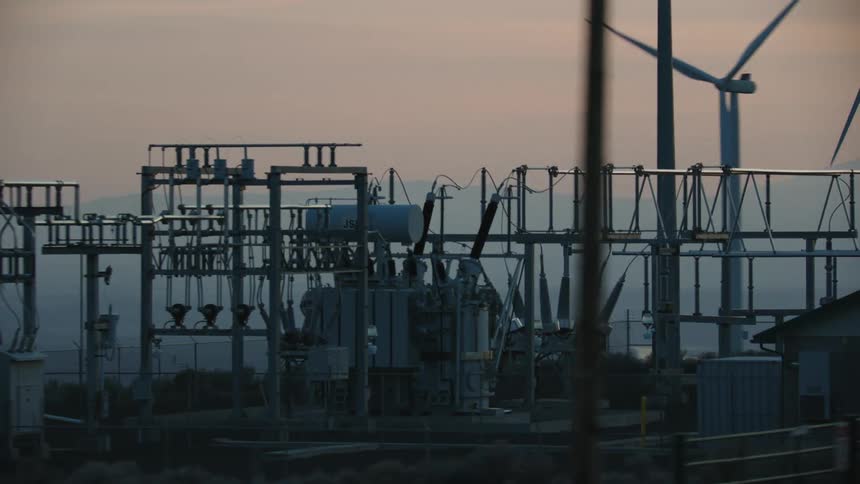 A power substation is seen with windmills and a large mountain in the background at sunset evoking the energy needs and evolution of energy in the world