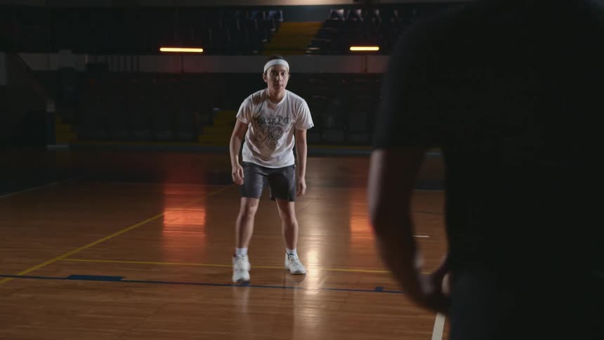 A man tossing a basketball to a girl and she dribbles the ball and passes it back in a gym with seats in the background