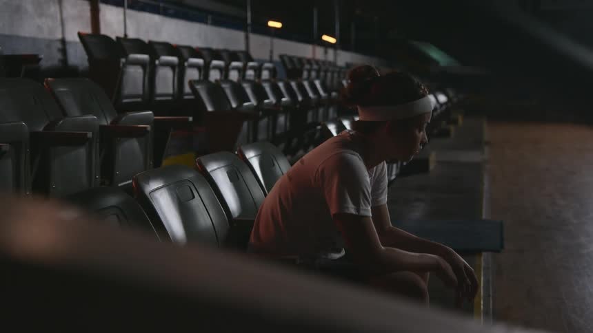 Teenager sits in spectator seating in dark room bending forward ang hanging her head