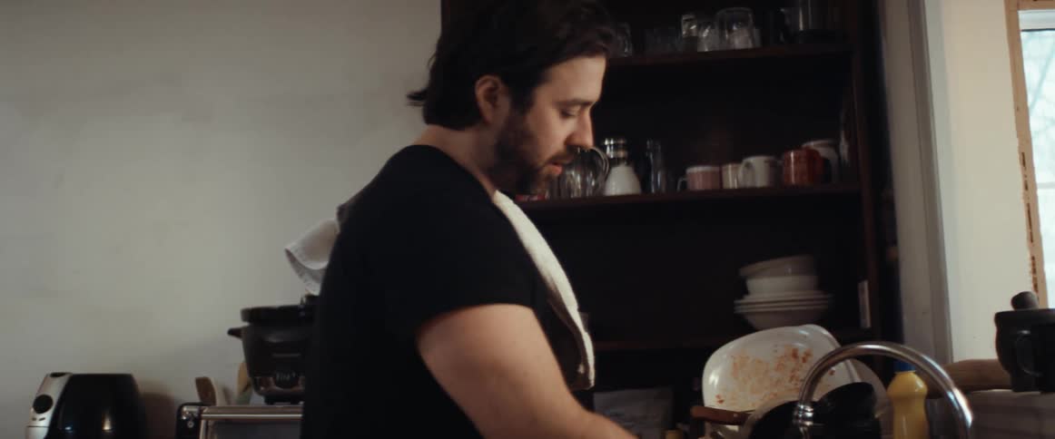 A man in a kitchen washing dishes with a towel over his shoulder