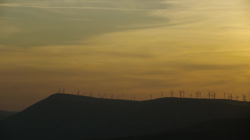 Wide shot in golden sunset light of a silhouetted ridge of wind turbines generating electricity for small businesses and residential customers