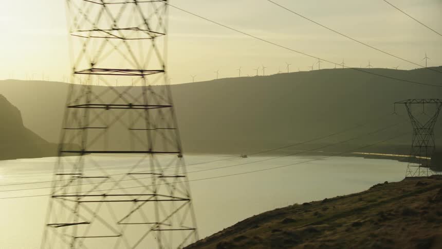Large transmission towers moving electricity across America are seen next to the Columbia river at sunset with Windmills on the ridge in the background