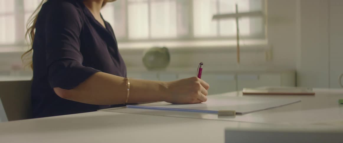 A woman sitting on a chair at a table drawing with a wall and window in the background