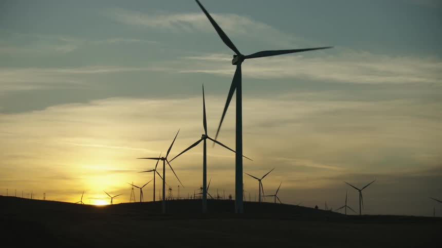 Many wind turbines rotate as the sun dips behind the horizon on a beautiful clear day as clean energy is being produced by the wind only