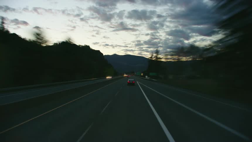 Time lapse blue hour shot as traffic flows along road bordered by forest covered mountain and river stopping at red traffic light after passing mcdonalds restaurant