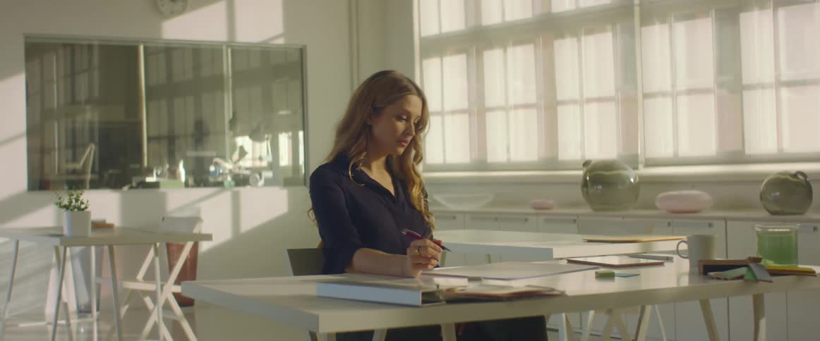 A woman sits at a large desk thinking and then starts writing on the paper in front of her
