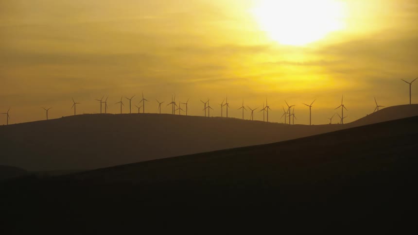 Low altitude aerial shot at sunset looking back at a ridge of silhouetted wind turbines generating clean green energy that is renewable