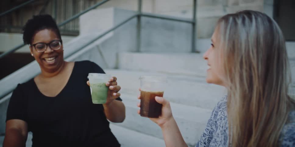 Two women sitting on a staircase with a banister in front of a building knocking cups together