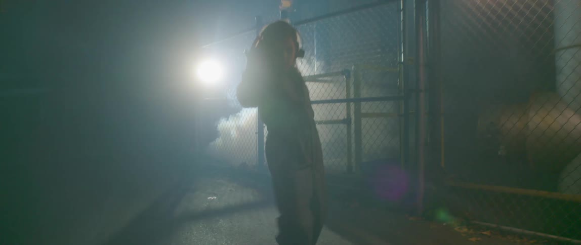 A woman wearing headphones and dancing near smoke and a chain link fence at night