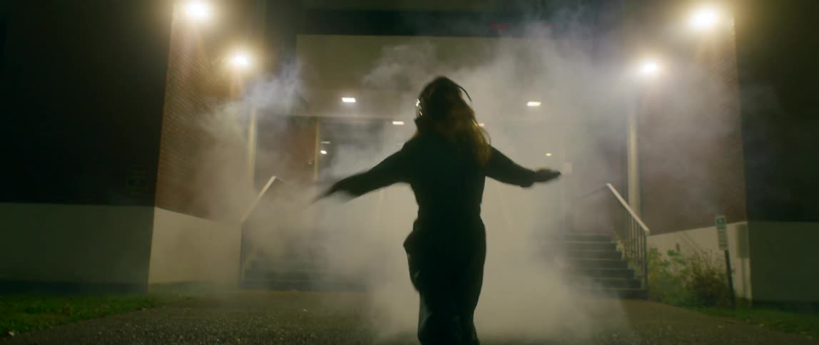 A woman dances erratically with aggressive fullarm movements at night in front of schoollike building as lights flash and smoke billows