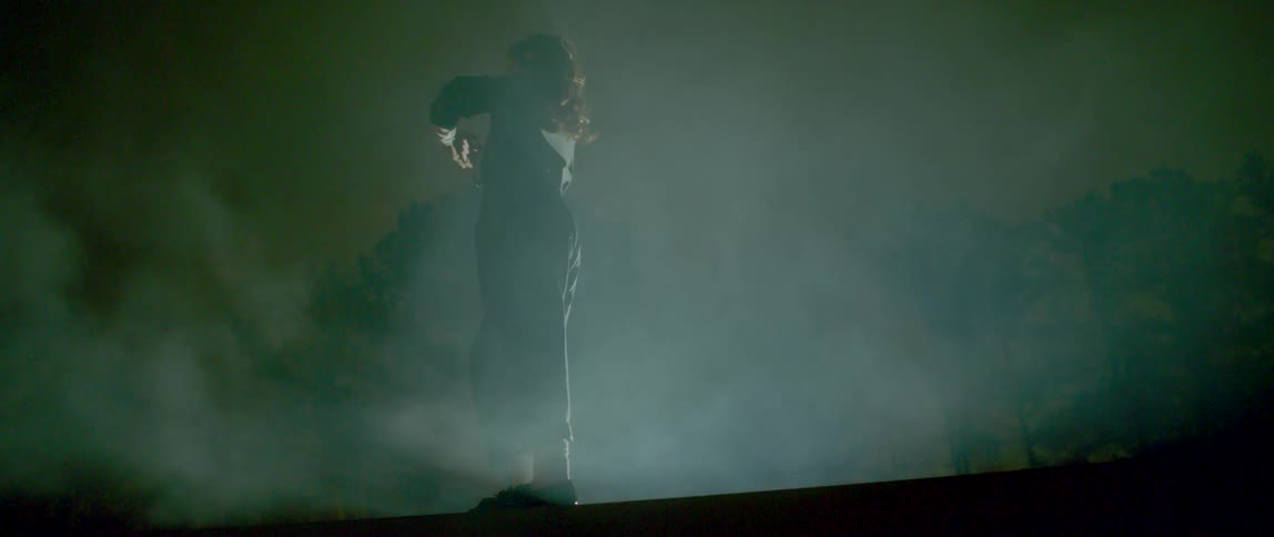 A woman dances on a ledge in a boiler suit as smoke rises around her