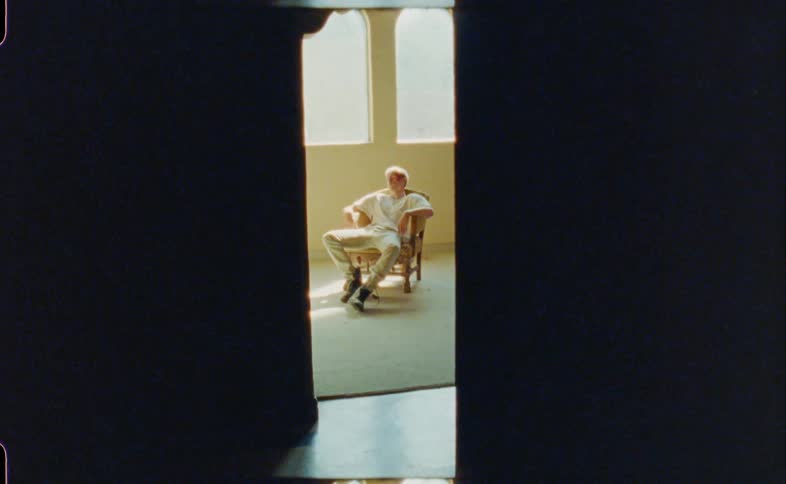 Blonde man with a white t shirt jeans and black boots sits in an armchair in a room
