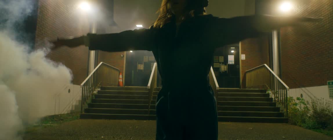 A woman in headphones dancing in front of a building with smoke in the background at night