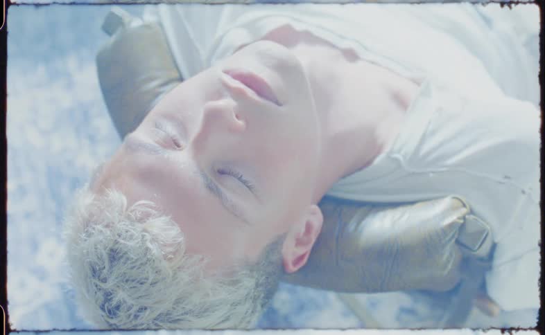 Blonde man with a white t shirt sits in a brown armchair eyes closed