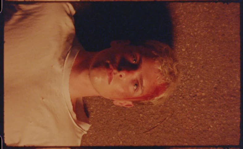 Blonde man lying on ground with blood on side of head abruptly opens eyes and sits up