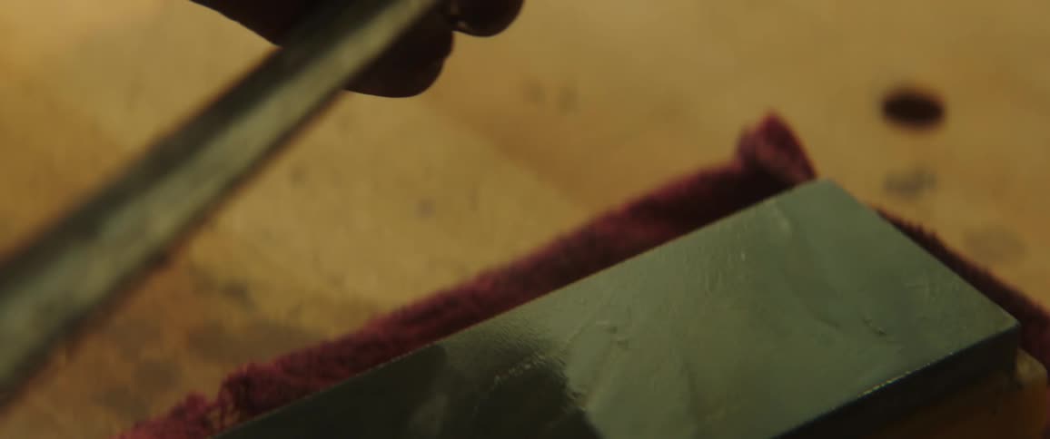 A person carefully sharpens a knife using a whetstone on a wooden crafting table