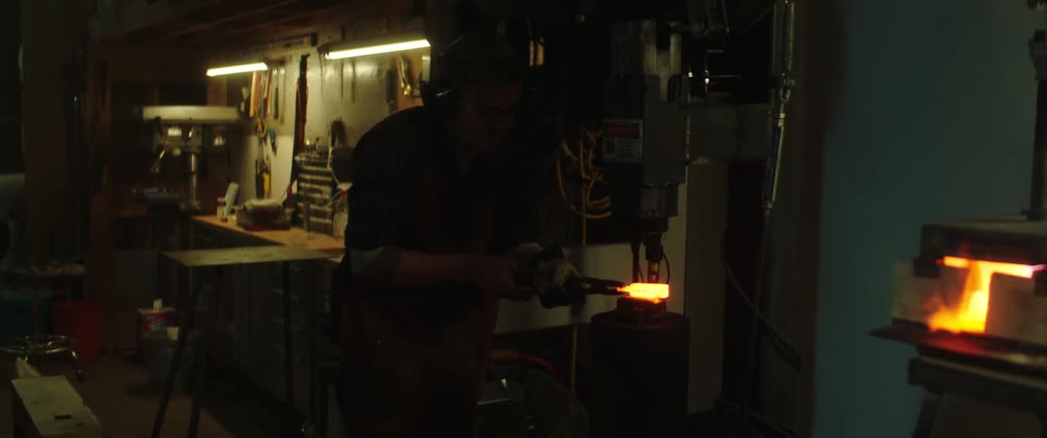A man uses a kiln to make a piece of metal glowing hot then places it under a press