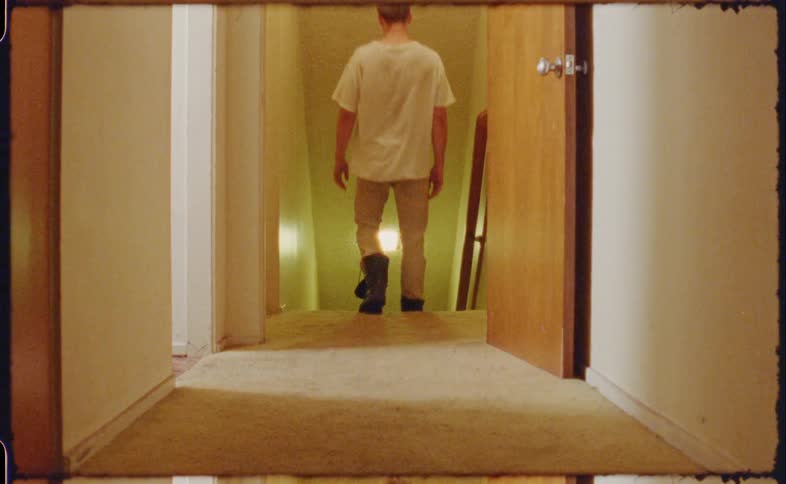 A male wearing boots walks out of a room and down a carpeted hallway before descending some stairs
