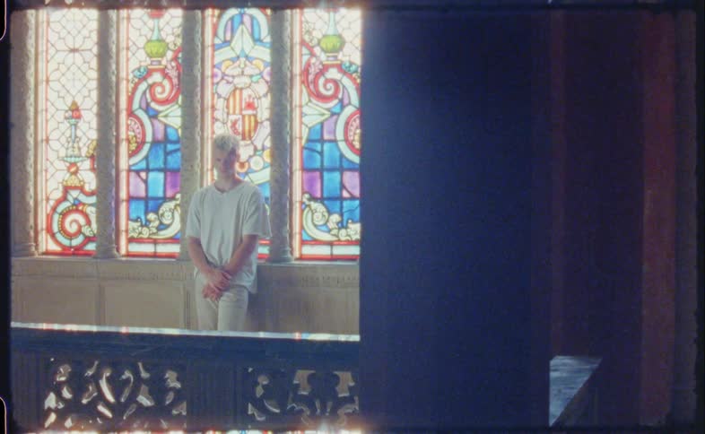 A man looking down standing up against a wall in front of a stained glass window in a chapel