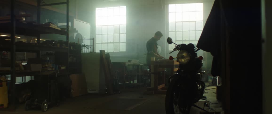 A silhouette of a man in a room with a motorcycle in the foreground and a wall and windows in the background