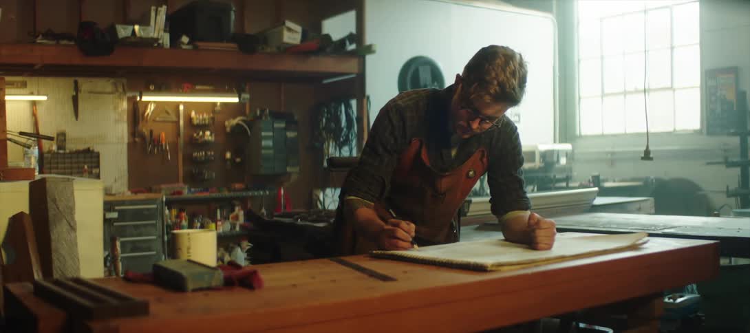 A man leans on a table in a workshop and writes on a large piece of paper