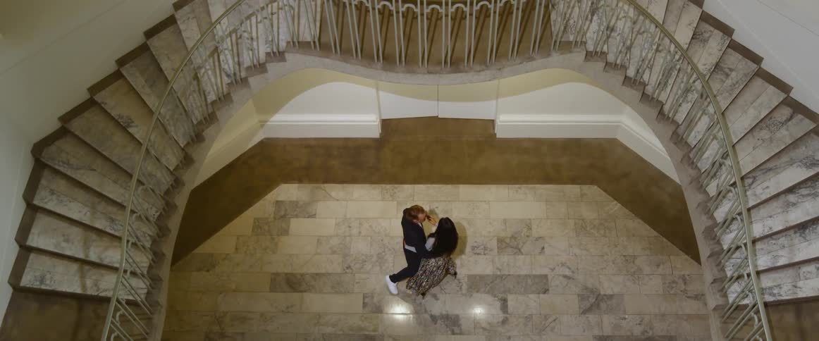 A man and man dance together in a large mansion by tall dual staircases