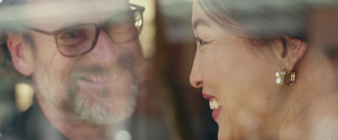 A man and woman laugh as they stand close together near a window