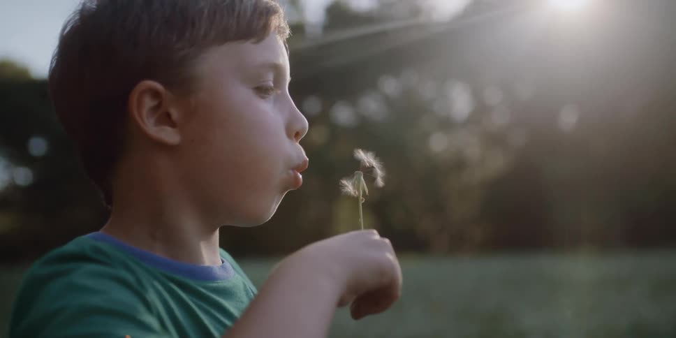 A child blowing the fuzz off a dandelion outside near a wooded area