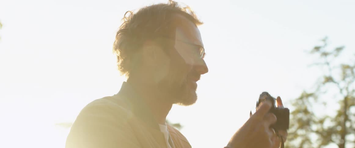 Middle aged man with glasses a beard and a cream sweater takes photographs with a camera with shining sun in the background
