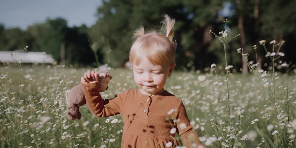 Blonde little girl with a brown dress holding toys walks in the field with flowers