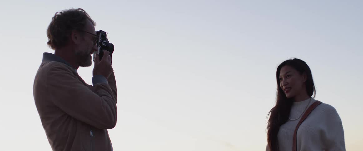 Middle aged man taking photographs of woman wearing a white sweater is silhouetted against the sky