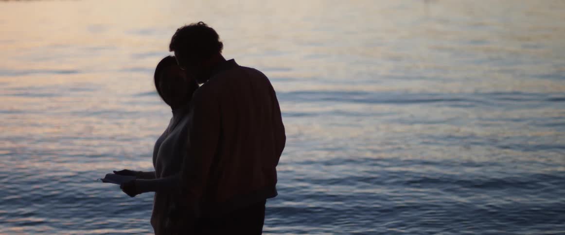A man walks up to a woman by the water and hugs her