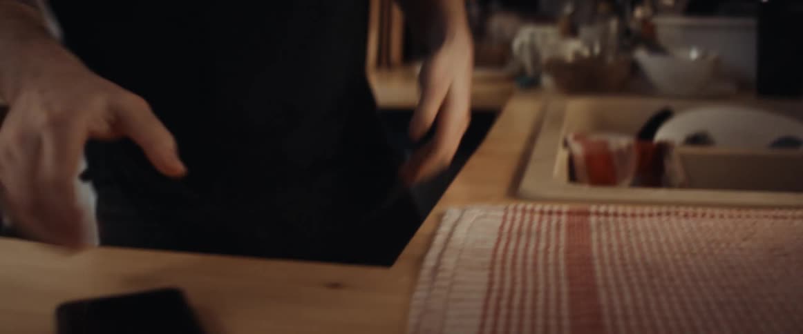 A man is drying dishes and picks up his cell phone and starts talking