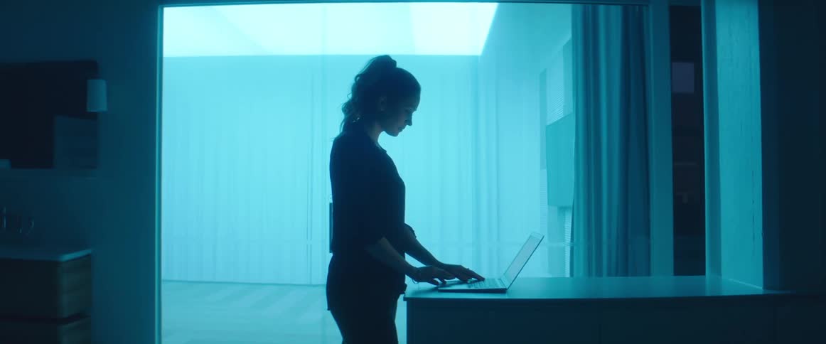 A woman in a room standing by a table and typing on a laptop with a wall in the background