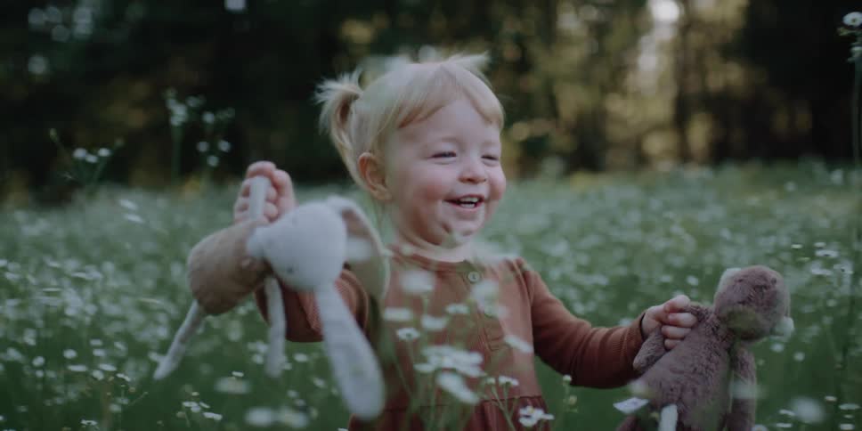 A little girl holding two stuffed animals smiling and running through a field of flowers in a forest