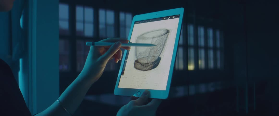 In a darkened room a woman holds a tablet with one hand and draws a sketch with a stylus in the other