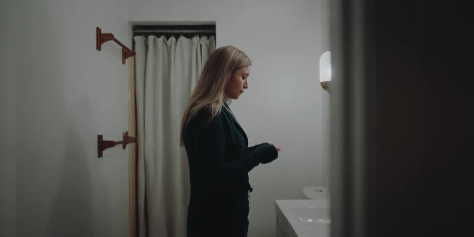 A woman in a bathroom with a nervous expression on her face picks up and checks a pregnancy test from off the sink counter