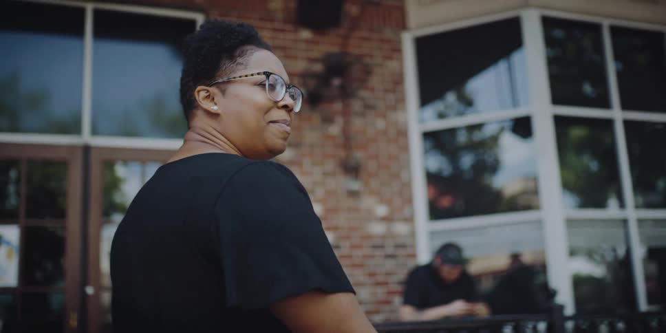 A woman with glasses walking while holding a cup with a building and a fence in the background