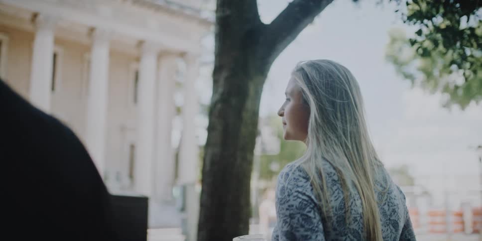 Woman walks along street with friend listening as they talk with large building with white columns in background