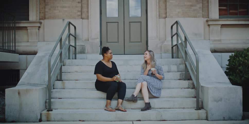 Two woman sitting on the steps of a stairway in front of a building drinking and laughing with each other