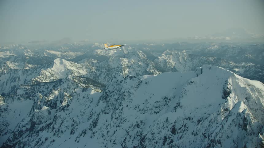 Orange and white single engine airplane flying straight and levels turns radically and sharply away from camera without warning revealing snow covered mountains in the background