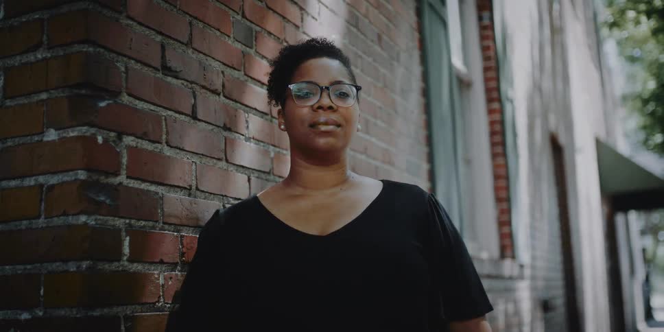 A woman standing near the brick facade of a building turns to look into the camera