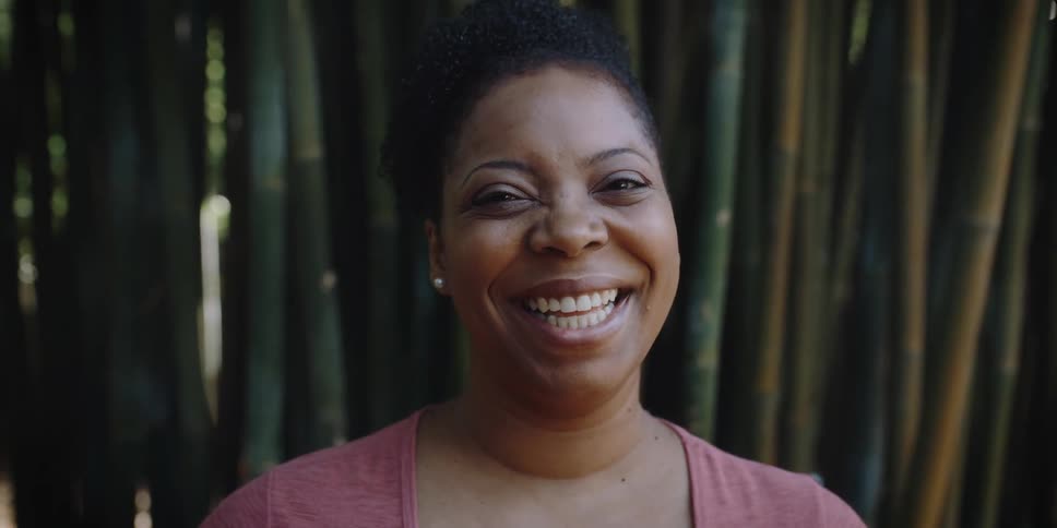 A black woman in a light red shirt smiles brightly and laughs in front of a shaded dense wall of bamboo