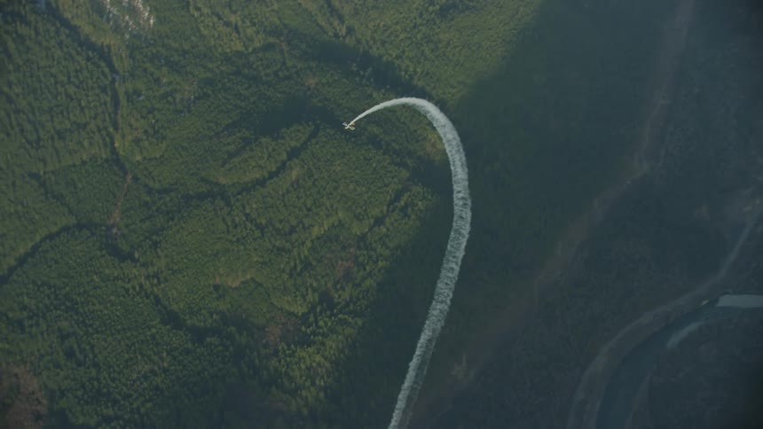 Airplane flies over river and forest partially in shadow and partially in sunlight with smoke as it climbs straight up at the camera doing a loop